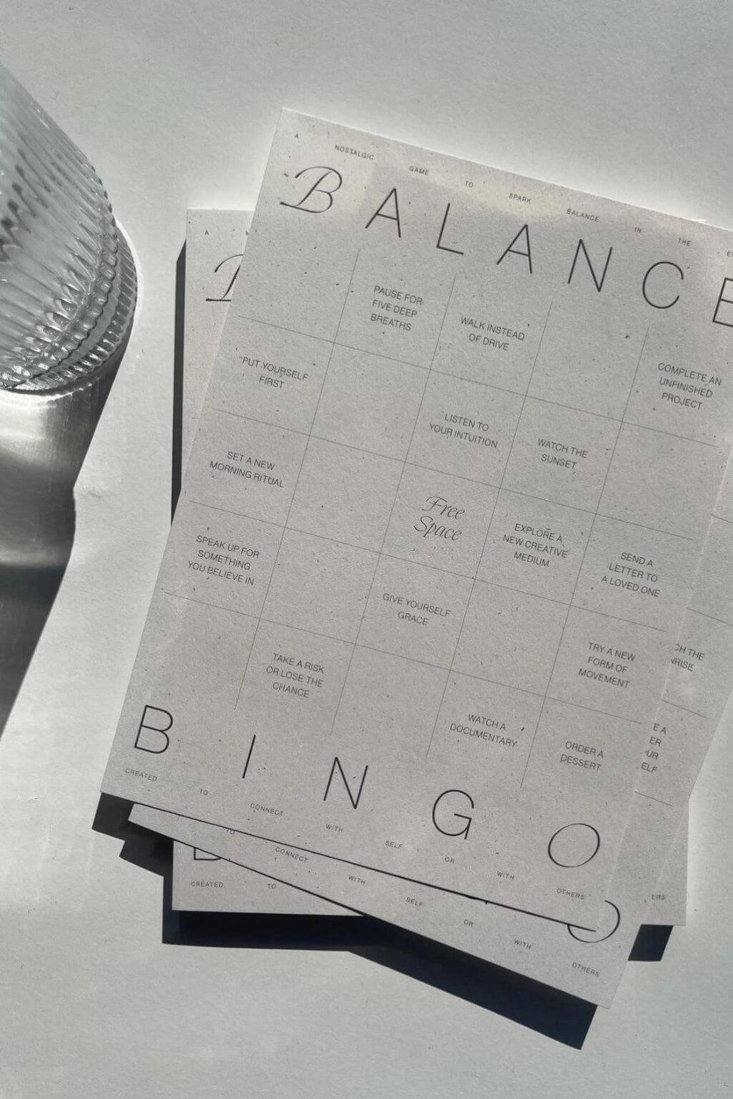 A stack of minimalist balance bingo cards with black text on a light gray background, styled on a light surface with a textured glass object partially visible on the left.