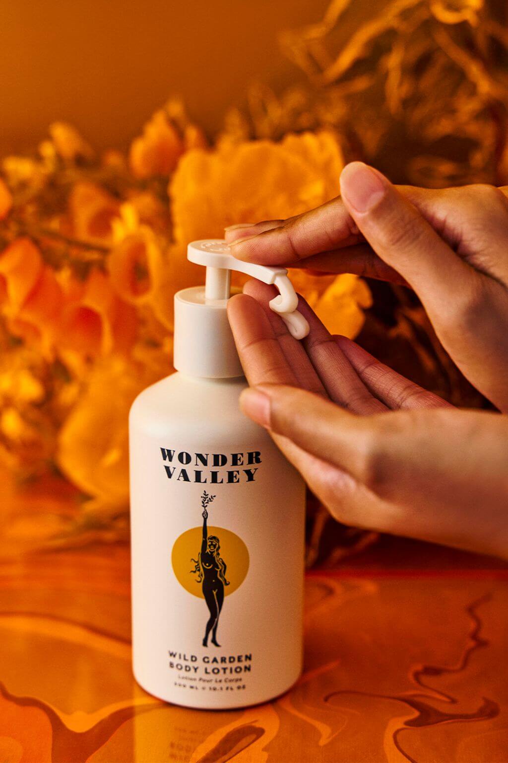 A person's hands are shown dispensing lotion from a white pump bottle labeled 'WONDER VALLEY WILD GARDEN BODY LOTION' against a warm orange background with dried flowers.