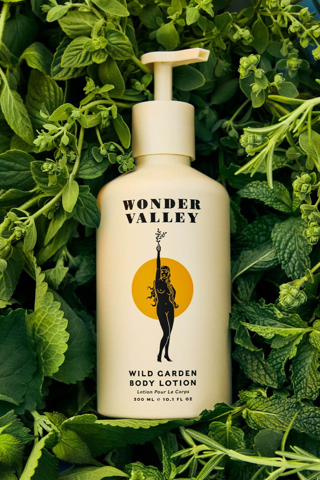A bottle of Wonder Valley Wild Garden Body Lotion placed among dense green leaves and herbs. The bottle is cream-colored with a pump dispenser and features a black silhouette logo with a yellow sun behind it.