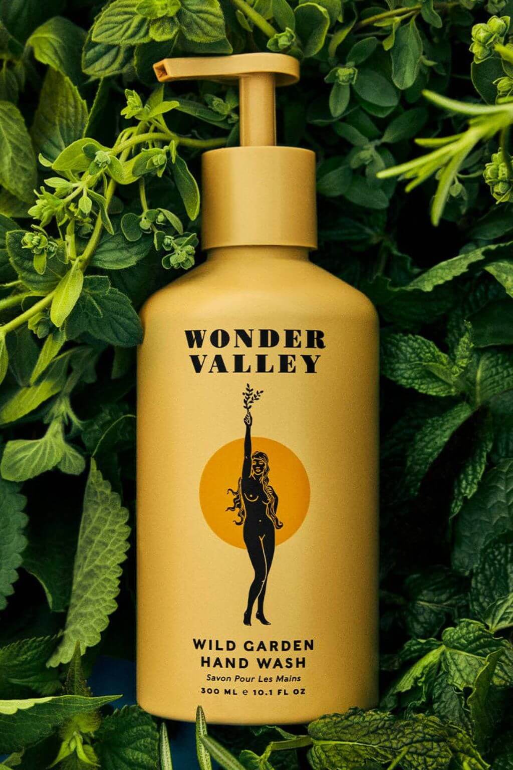Wonder Valley Wild Garden Hand Wash in a matte mustard-yellow bottle with a matching pump dispenser, surrounded by lush green leaves and herbs. The label features black text and a silhouette graphic.