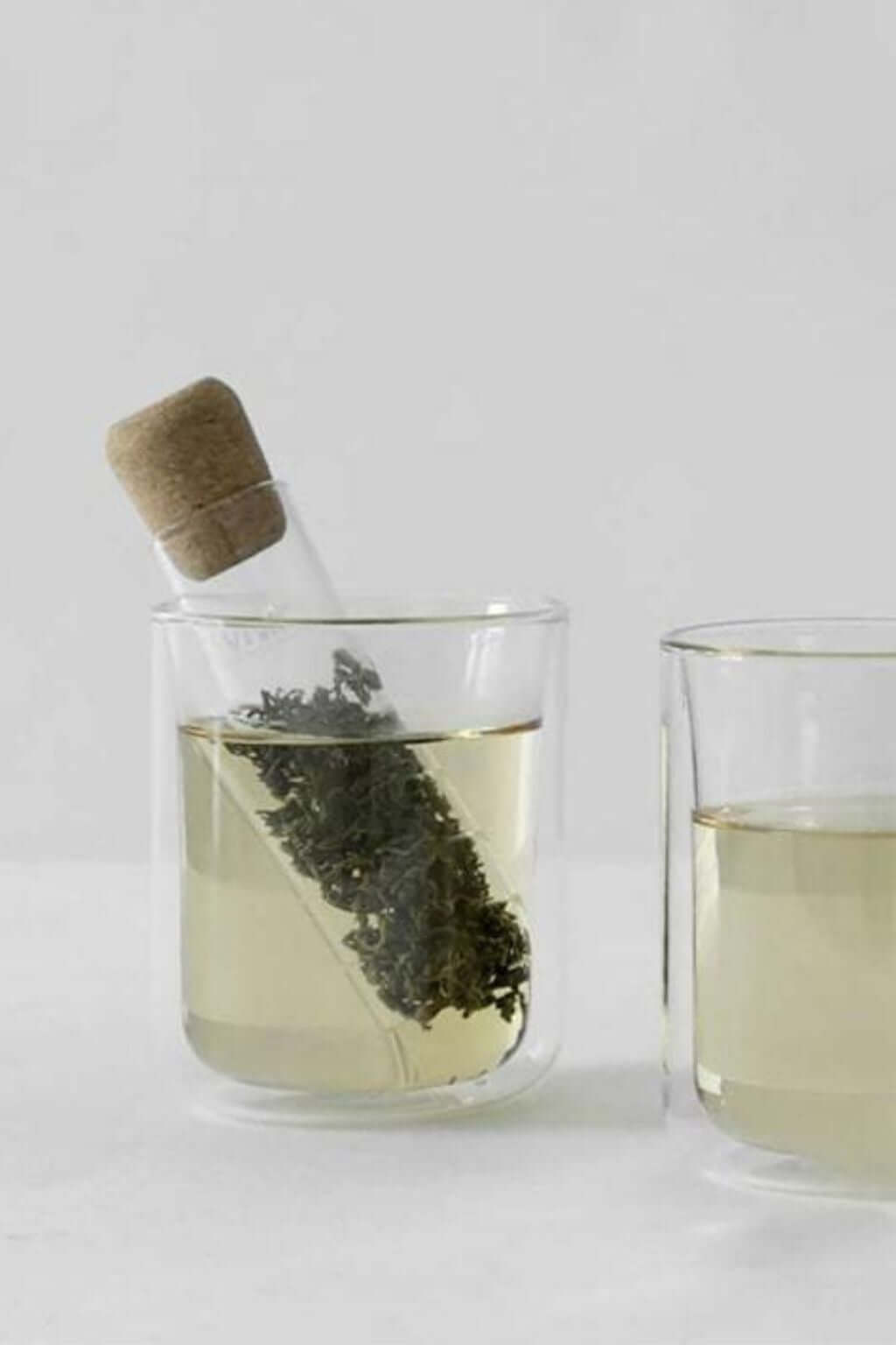 A clear glass tea infuser tube with a cork stopper filled with tea leaves, alongside two empty glass cups.