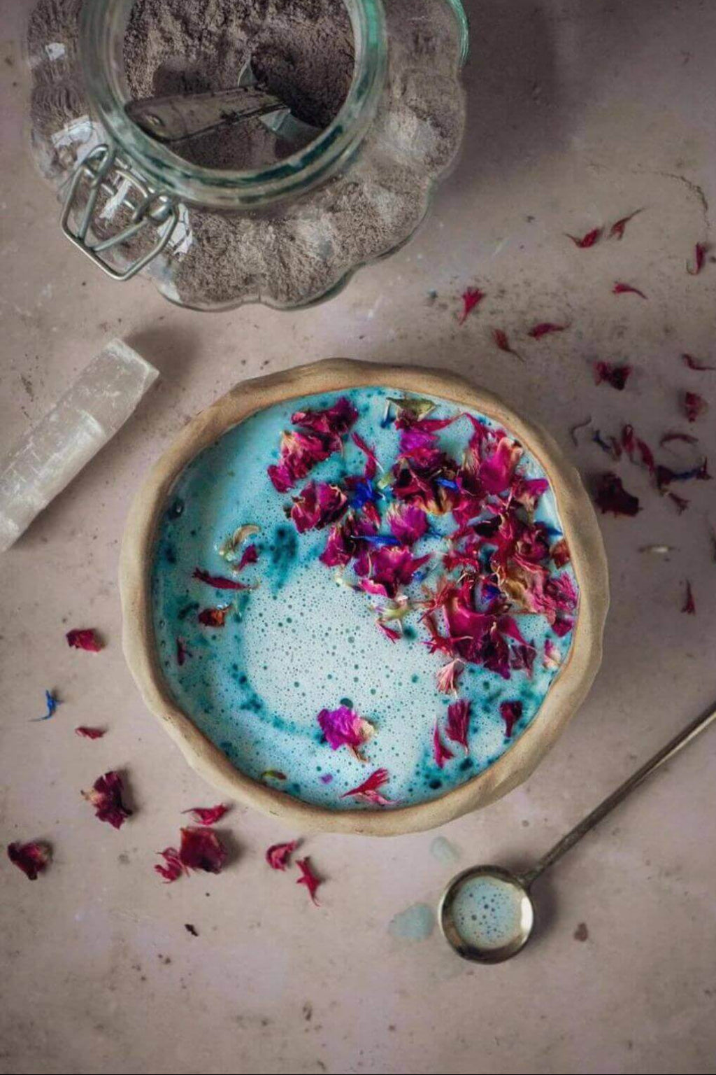 A ceramic cup filled with bright blue beverage topped with dried pink and red flower petals, arranged on a neutral surface with a glass jar of powder, a brass spoon, and scattered petals.