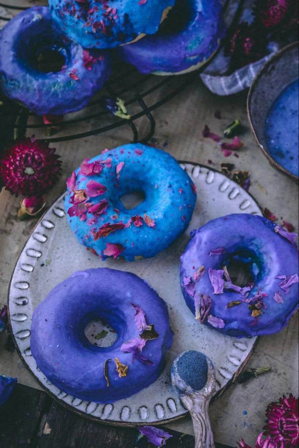 Colorful glazed donuts with blue and purple frosting, decorated with dried flower petals, arranged on a white scalloped plate with a spoon, set on a rustic wooden table with flowers.