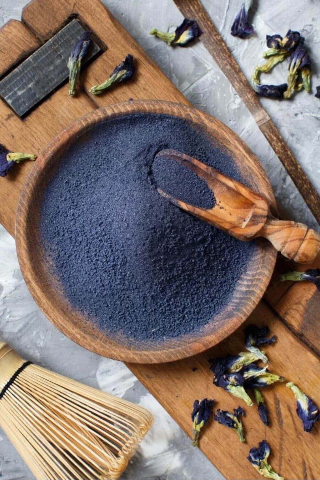 A wooden bowl filled with dark blue butterfly pea powder, with a wooden scoop resting inside, photographed from above on a rustic wooden board with dried flower petals and a bamboo whisk nearby.