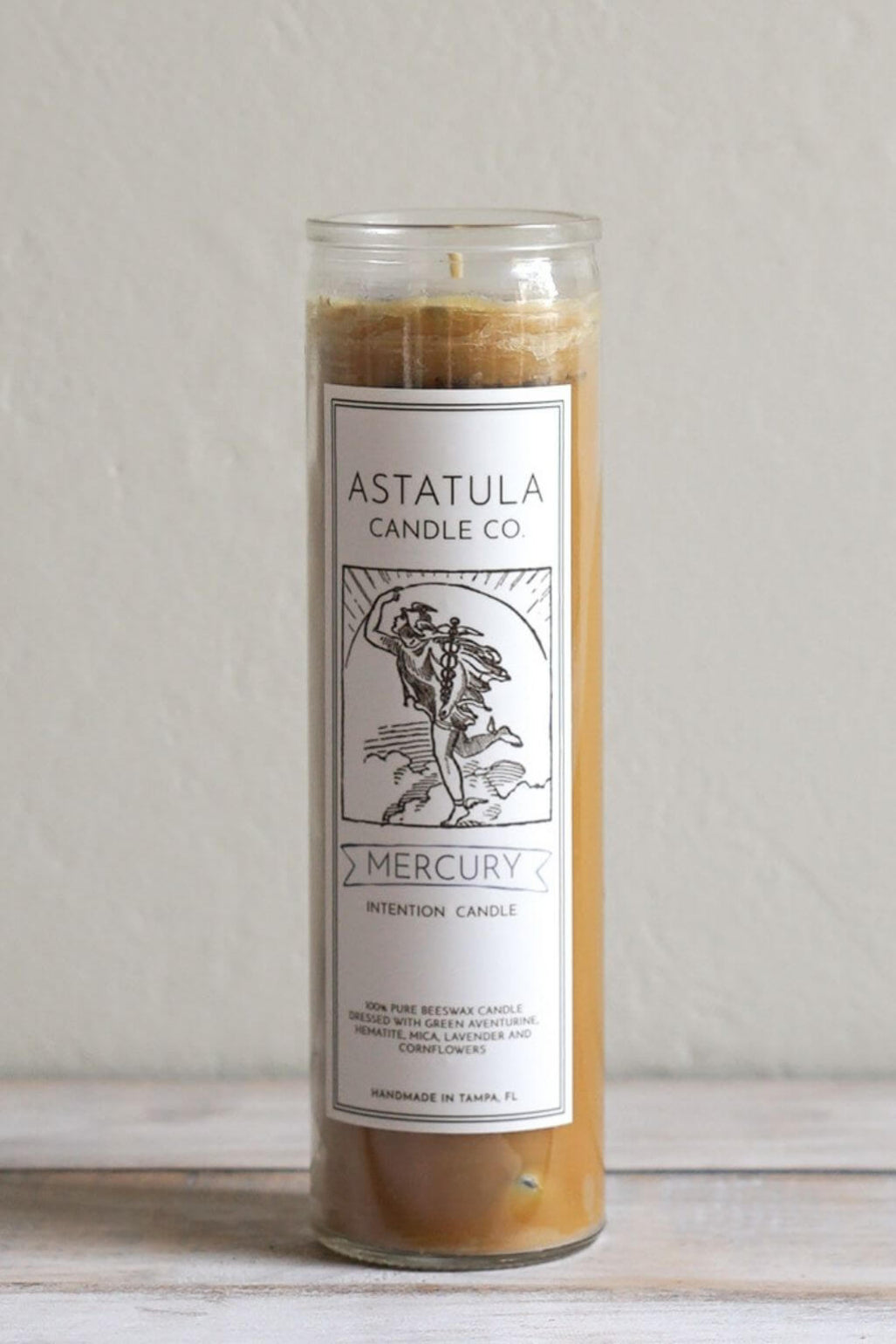 A tall glass jar candle with a white label reading 'ASTATULA CANDLE CO' and 'MERCURY', filled with warm tan wax, standing on a light surface against a plain light gray wall.