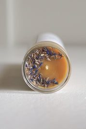 A close-up of a clear glass jar showing the top of a candle with a warm beeswax surface and scattered dried lavender buds.