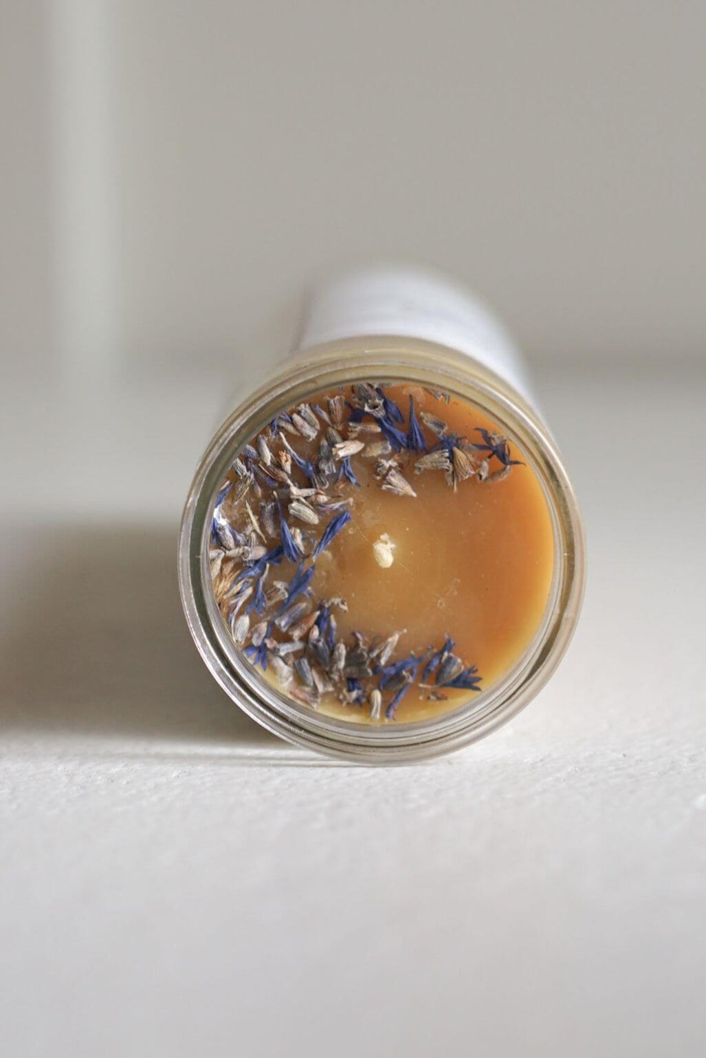 A close-up of a clear glass jar showing the top of a candle with a warm beeswax surface and scattered dried lavender buds.