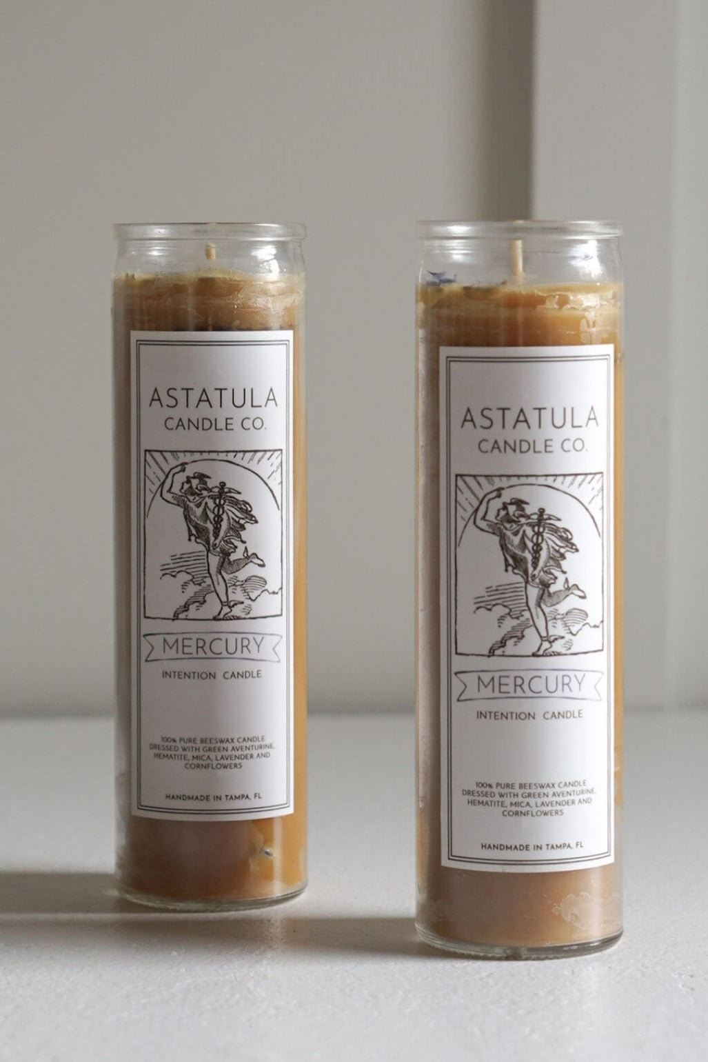 Two tall glass jar candles with white labels reading 'ASTATULA CANDLE CO' and 'MERCURY' are shown on a light neutral surface against a soft gray background.
