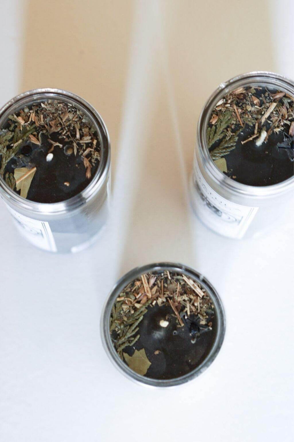 Three dark beeswax candles with dried botanicals and wood shavings, arranged on a light surface.