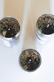 Three dark beeswax candles with dried botanicals and wood shavings, arranged on a light surface.