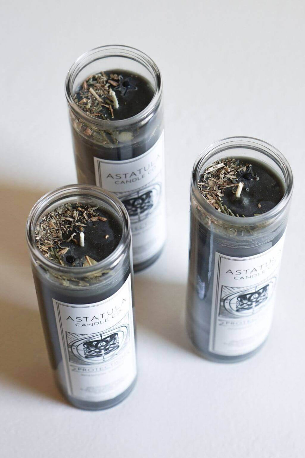 Three glass jar candles filled with dark wax and dried botanicals, arranged on a light neutral background. The labels read ASTATULA CANDLE CO.