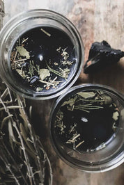 Two clear glass jars filled with dark beeswax topped with dried leaves and botanicals, arranged on a rustic wooden surface with a bundle of dried herbs and a dark stone nearby.