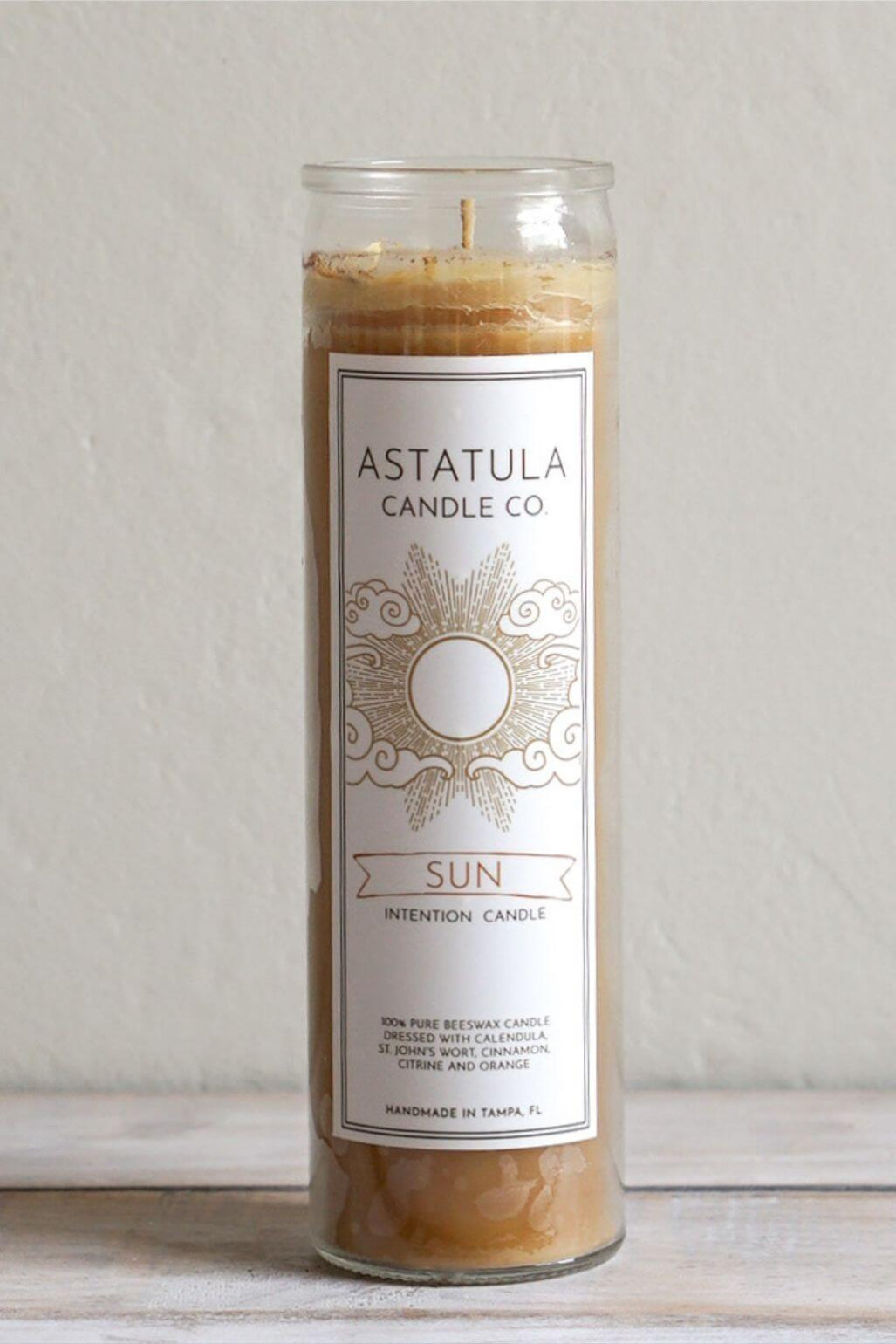 A tall glass jar candle with a white label reading 'ASTATULA CANDLE CO' and 'SUN INTENTION CANDLE,' filled with warm tan beeswax, standing on a light surface against a neutral wall.