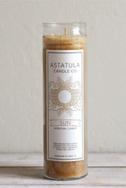 A tall glass jar candle with a white label reading 'ASTATULA CANDLE CO' and 'SUN INTENTION CANDLE,' filled with warm tan beeswax, standing on a light surface against a neutral wall.