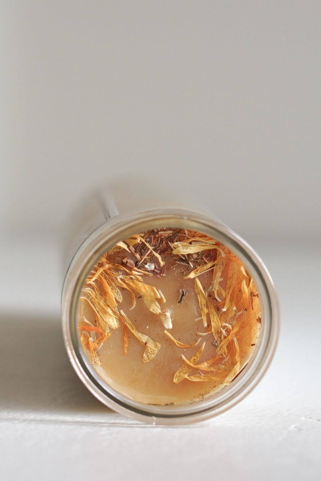 A clear cylindrical glass candle viewed from above, containing light beeswax with dried tan and brown botanical pieces on top, set against a plain light background.