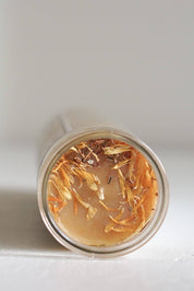 A clear cylindrical glass candle viewed from above, containing light beeswax with dried tan and brown botanical pieces on top, set against a plain light background.