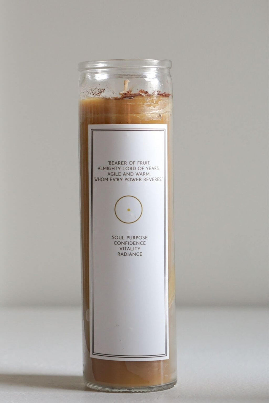 A tall glass jar candle with a white label, filled with light brown beeswax and topped with a small amount of brown and tan herbs. The label reads 'BEARER OF FRUIT' and 'SOUL PURPOSE CONFIDENCE WITNESS RADIANCE'.