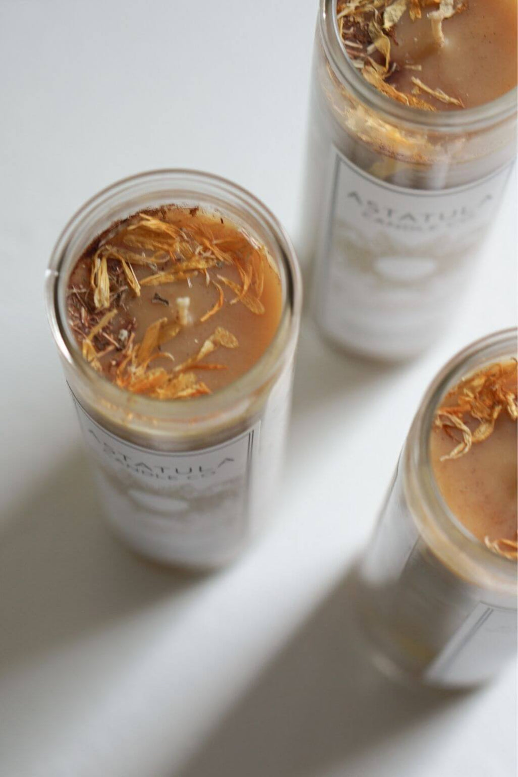 Three clear glass jars of Astatula Candle with white labels and dried botanicals on a light beeswax, arranged on a white surface.