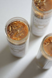 Three clear glass jars of Astatula Candle with white labels and dried botanicals on a light beeswax, arranged on a white surface.