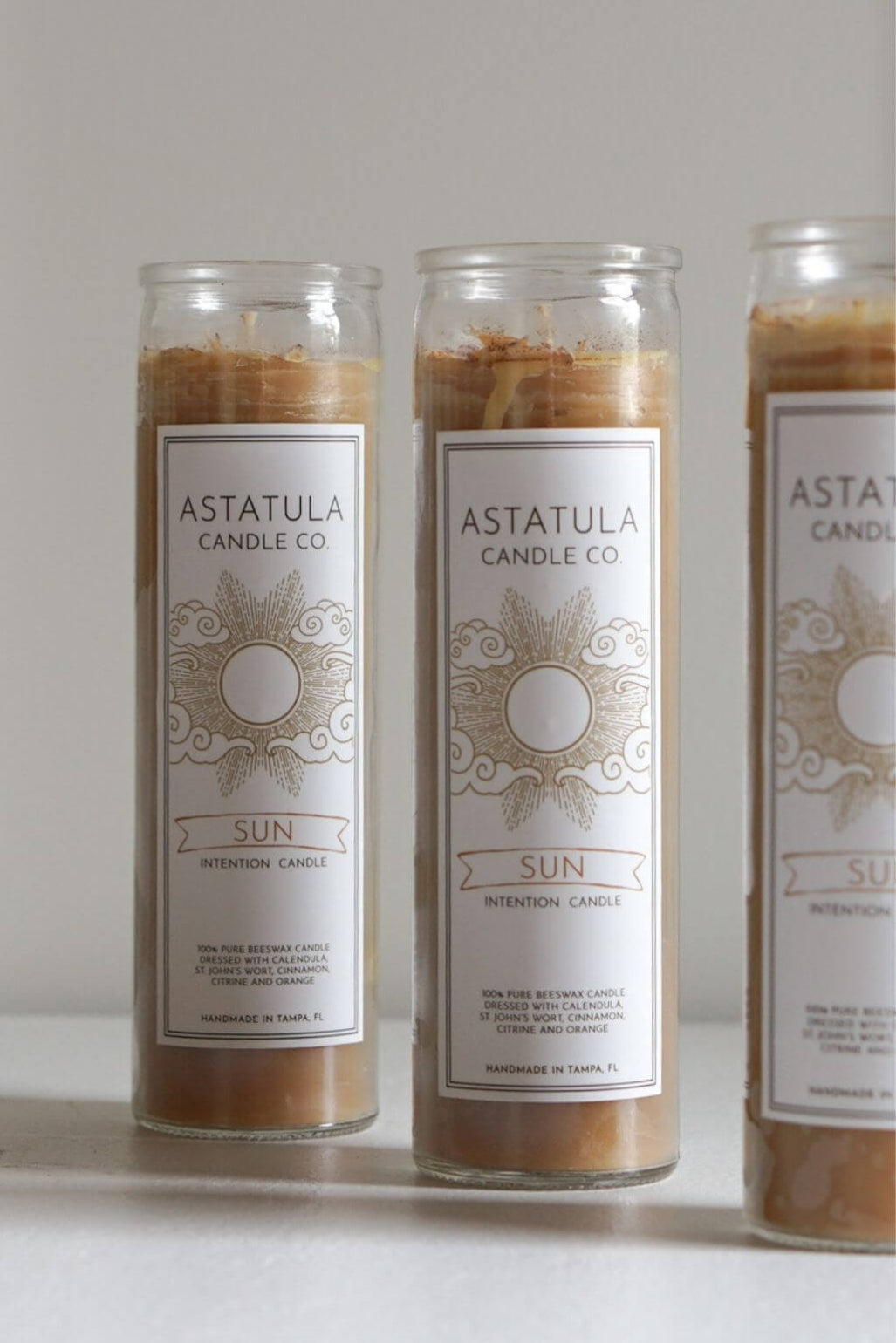 Three tall glass jar candles with white labels reading ASTATULA CANDLE CO. and SUN, filled with warm tan wax, arranged on a light surface against a neutral light gray background.