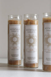 Three tall glass jar candles with white labels reading ASTATULA CANDLE CO. and SUN, filled with warm tan wax, arranged on a light surface against a neutral light gray background.