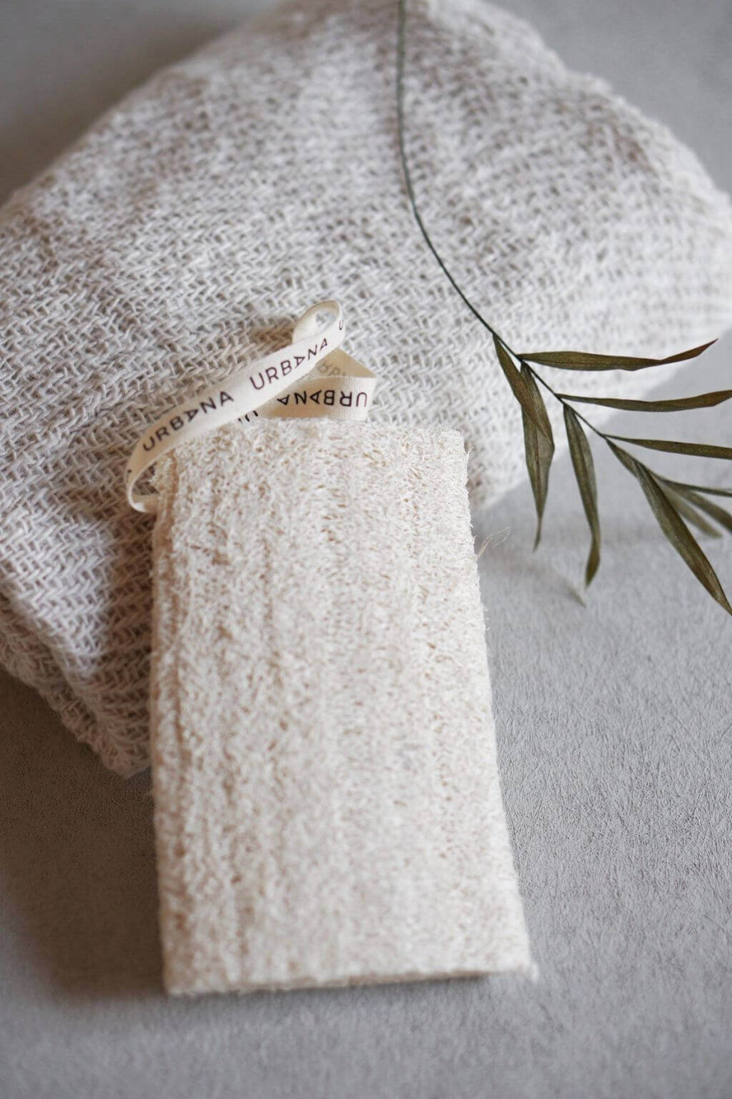 An off-white textured  sponge attached to a cream ribbon tag printed with 'URBANA' resting on a matching woven fabric background with a dried olive branch sprig.