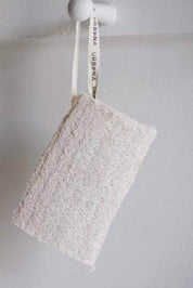 A rectangular natural loofah sponge with a white hanging loop printed with the brand name 'URBANA', suspended against a plain light gray wall.