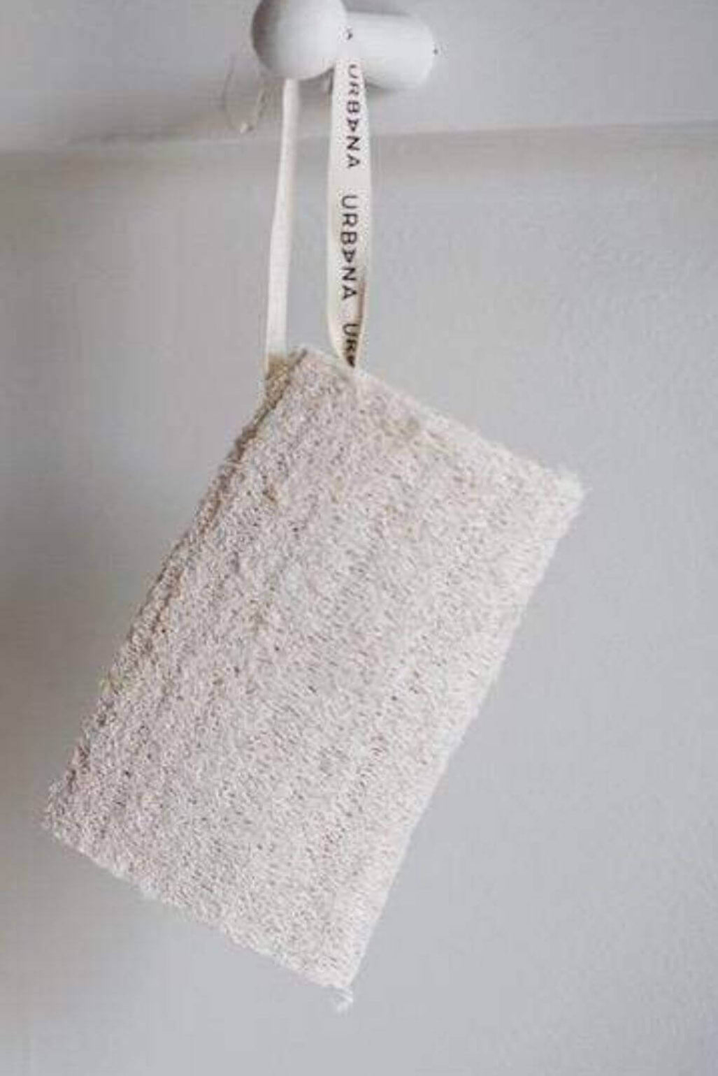 A rectangular natural loofah sponge with a white hanging loop printed with the brand name 'URBANA', suspended against a plain light gray wall.