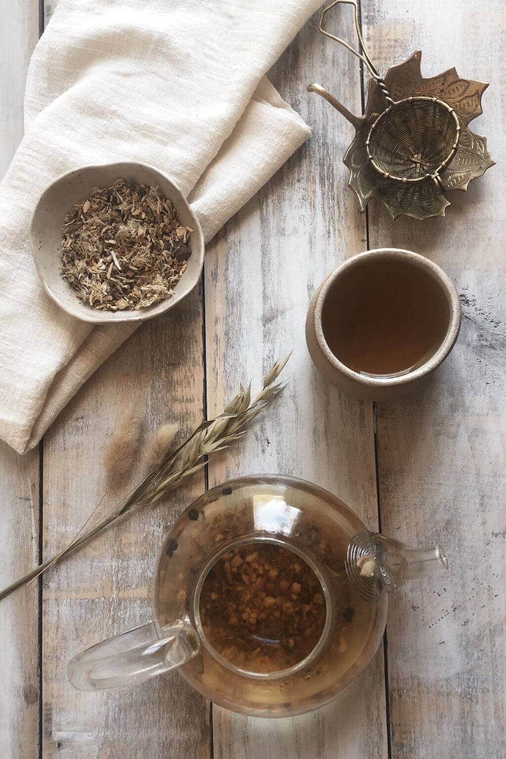 Brass tea strainer resting beside a handmade ceramic mug and glass teapot full of freshly steeped tea