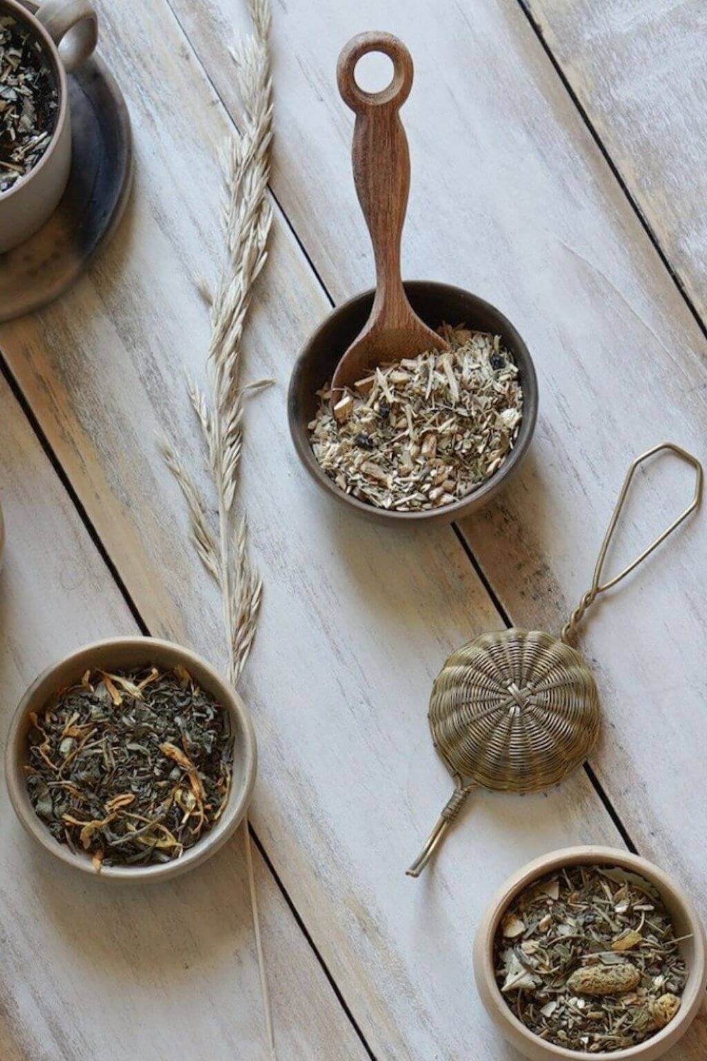 Artisan brass tea strainer upside down on a table beside several varieties of loose leaf herbal tea