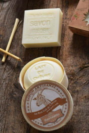 A styled product shot of Savonitto artisanal soap on a rustic wooden surface. The image features an open round tin of 'Lait de Chèvre' (cow's milk) soap showing its pale yellow contents, and the round lid in the foreground with a brown and cream label depicting a goat and the Savonitto brand name.