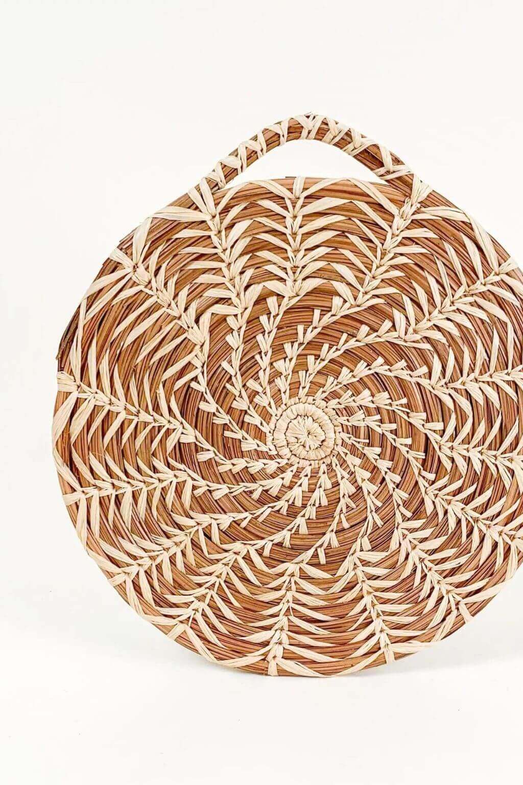 A round woven trivet with a single top handle, featuring a spiral pattern of natural brown and beige raffia on a white background.