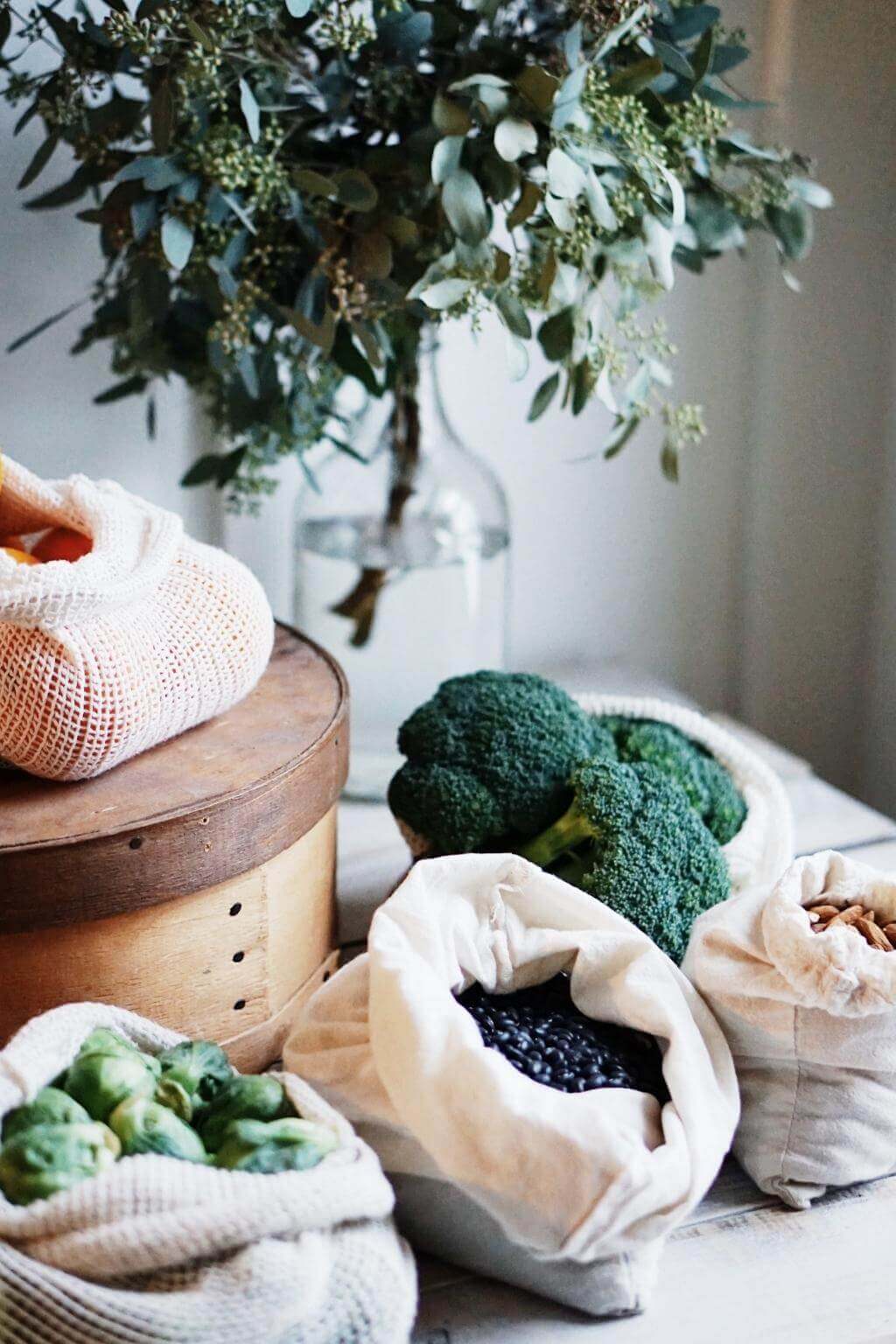 Broccoli and other vegetables in eco-friendly bags with a vase of greenery in the background.