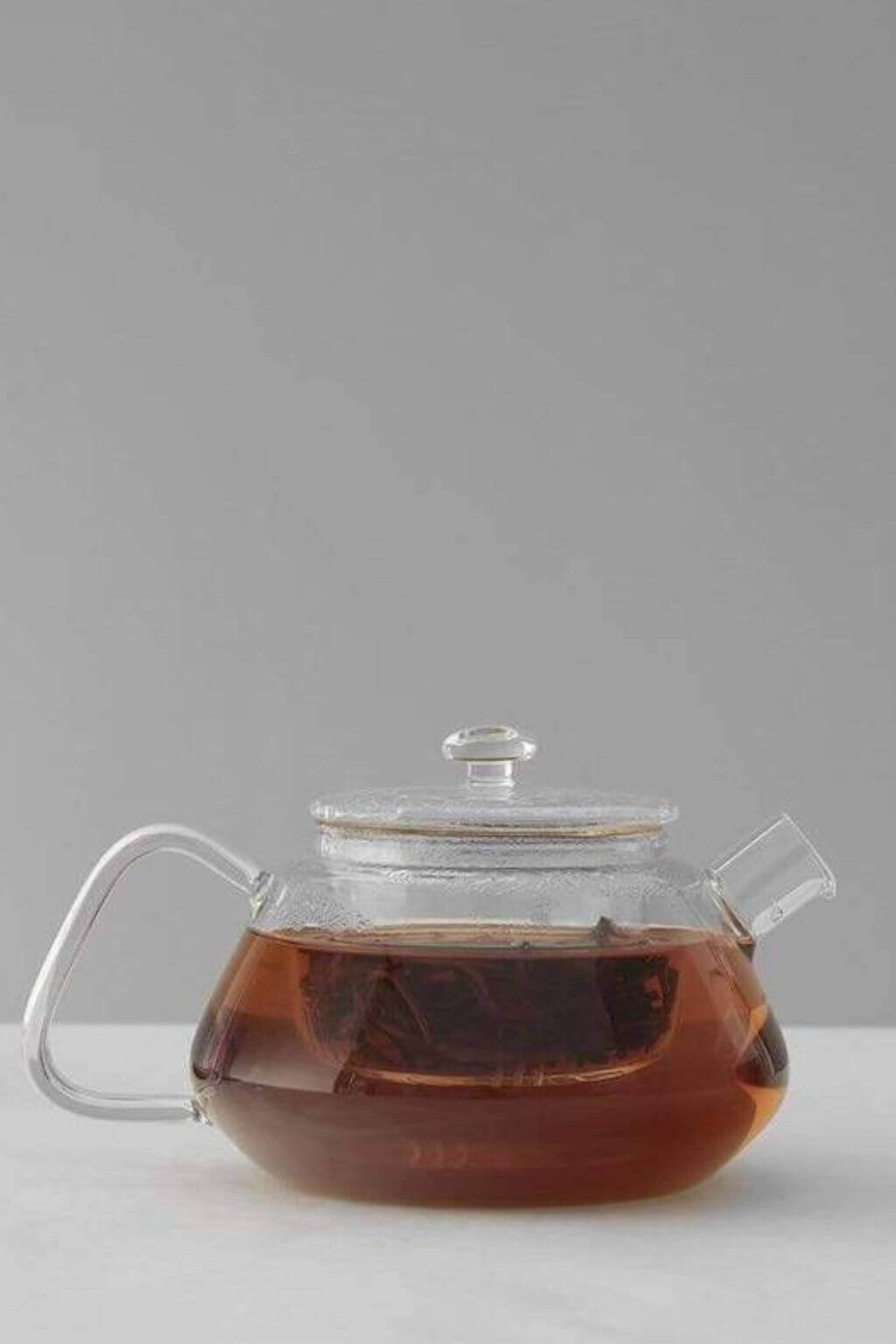 All glass teapot with a lid filled with tea on a light gray background