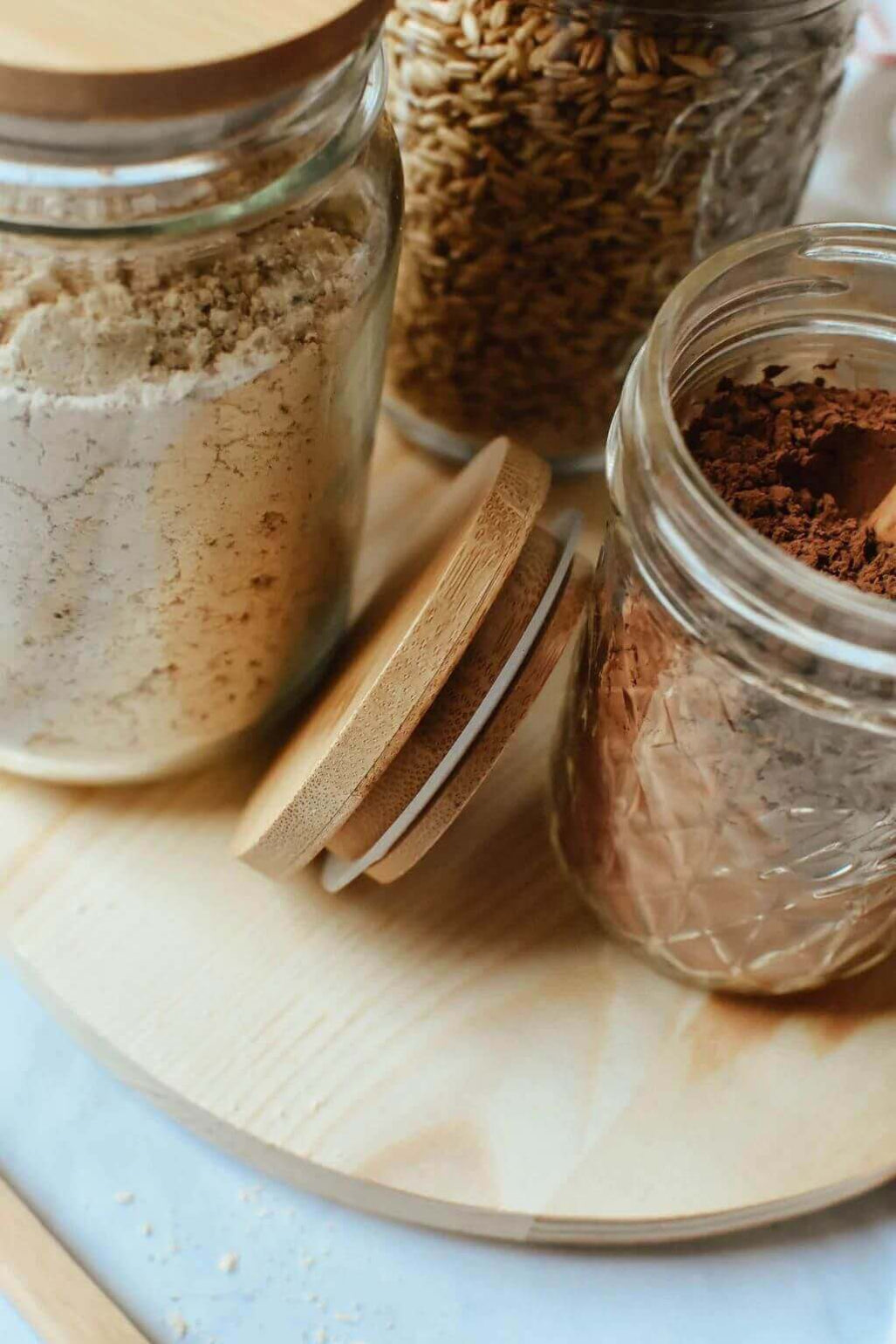 Clear glass storage jars with wooden lids containing many different things all arranged on a round wooden board.