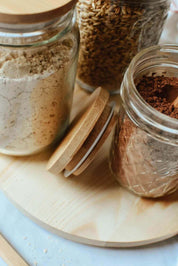 Clear glass storage jars with bamboo lids containing different things all arranged on a round wooden board.
